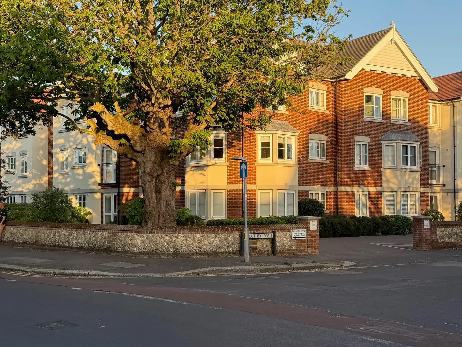 A red-brick apartment block with bay windows, set behind a mature tree on a corner plot