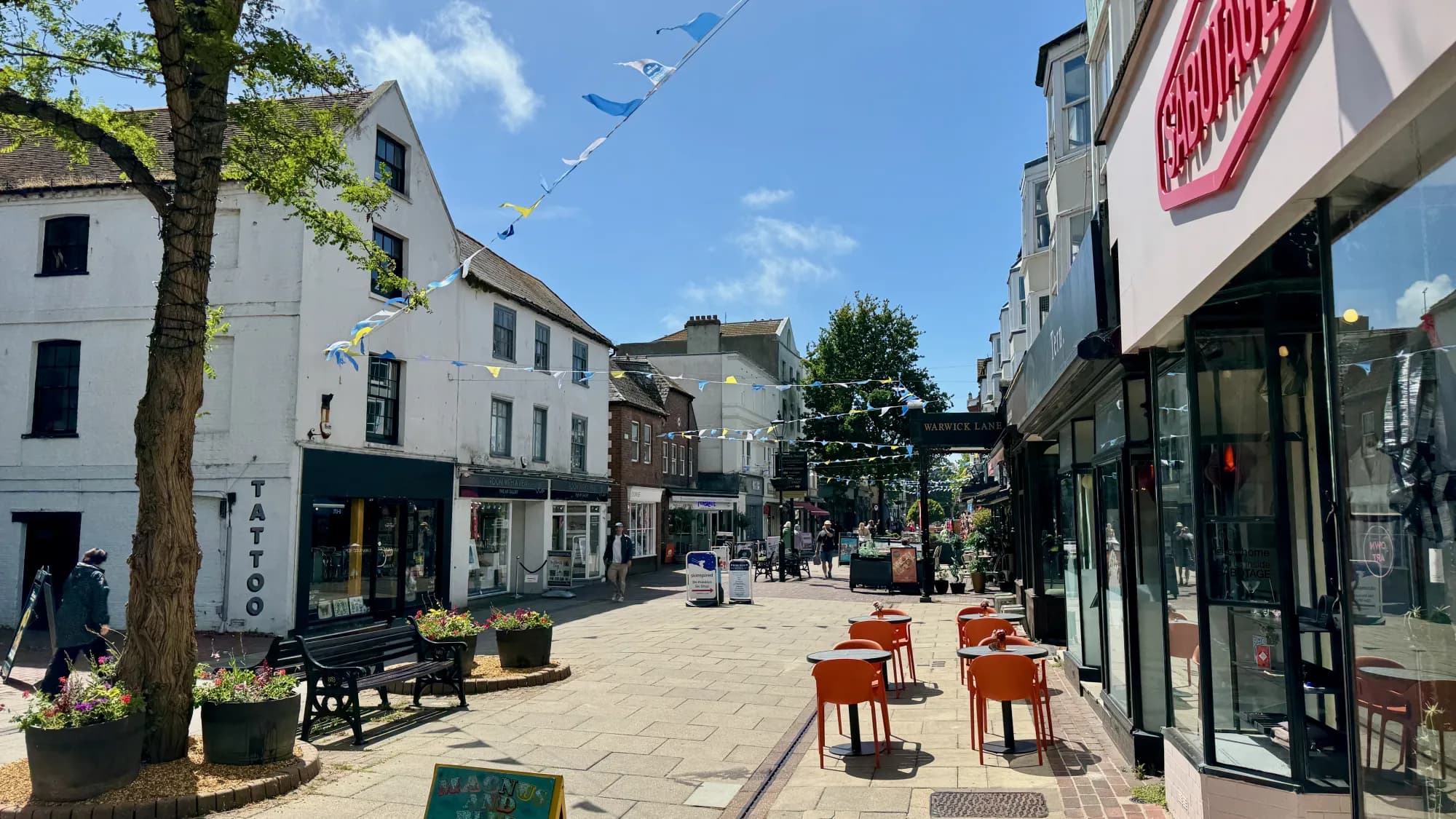 A pedestrianised street with benches, planters and café seating beneath a line of bunting